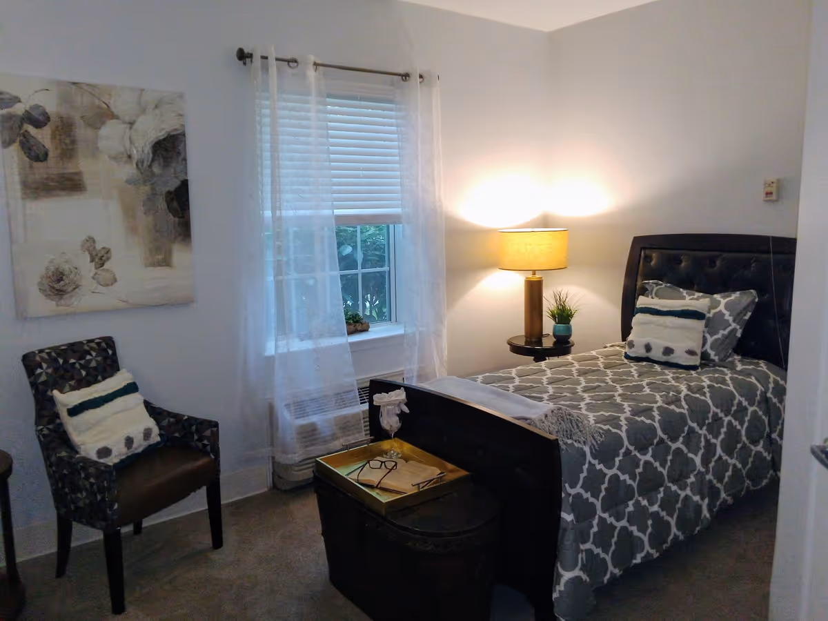 A cozy bedroom with a single bed featuring a patterned gray and white bedspread and two pillows. Next to the bed is a small round table with a lamp and a small plant. A window with white blinds and sheer curtains lets in natural light. A patterned armchair with a cushion is placed near the window, and a decorative painting hangs on the wall above the chair. A dark wooden chest at the foot of the bed holds a tray with glasses and a book.
