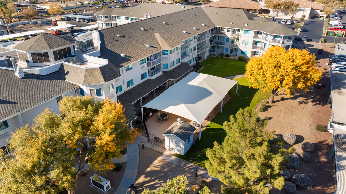 Aerial view of Vista del Rio senior living community showing a large multi-story building with a gray roof surrounding a green courtyard with a white canopy-covered seating area, trees with autumn foliage, and a parking lot in the background.