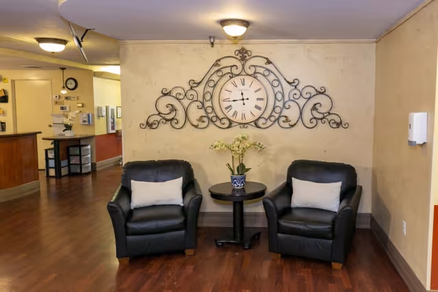 Two black leather armchairs with light-colored pillows are positioned on either side of a small round table with a potted plant. Above the table on the wall is a large decorative clock with ornate metal scrollwork. The room has wood flooring and beige walls, with a hallway and reception area visible in the background.