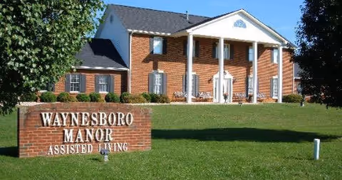 Front exterior view of a two-story brick building with white columns and a triangular pediment, surrounded by green grass and trees. A brick sign in the foreground reads 'Waynesboro Manor Assisted Living'.