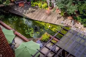 A serene outdoor garden area with a wooden deck surrounding a pond filled with lily pads and aquatic plants. Green umbrellas provide shade over seating areas, and lush greenery surrounds the space, creating a peaceful atmosphere.
