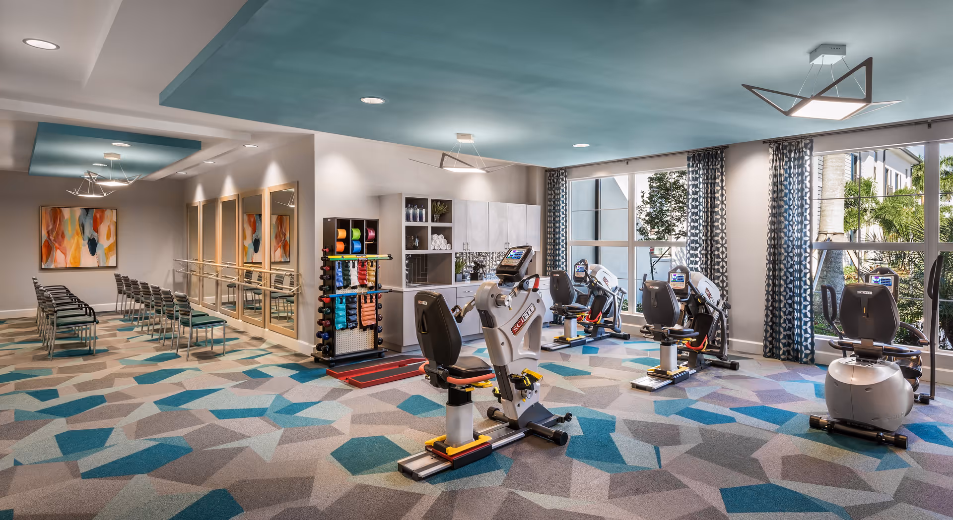 Bright assisted-living fitness room with exercise bikes, a rack of weights and equipment, stacked chairs, and large windows.