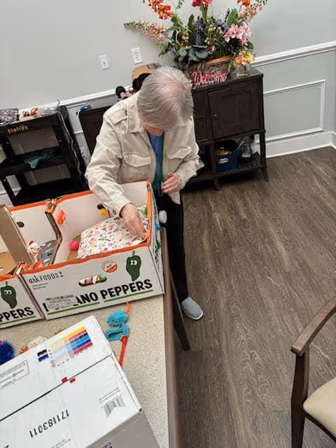 An elderly person with gray hair is standing next to a table with cardboard boxes labeled 'HABANERO PEPPERS' and other items on it. The person is looking down and appears to be organizing or handling something inside one of the boxes. The room has wood flooring, a dark wooden cabinet with a floral arrangement on top, and light-colored walls with white trim.