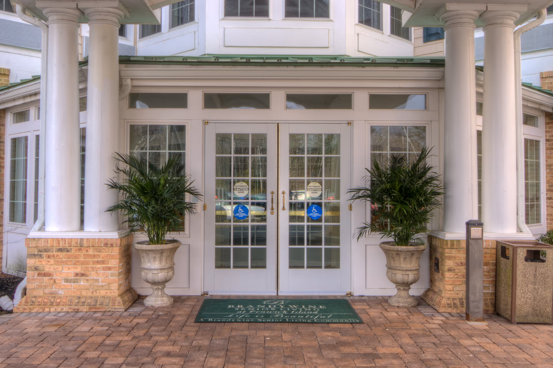Front entrance of a senior living facility with double glass doors framed by white columns, potted plants, and a welcome mat.