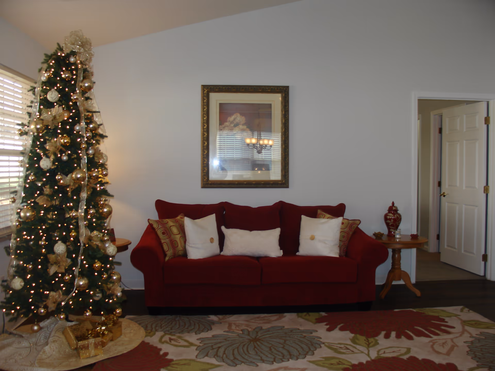 A cozy living room with a decorated Christmas tree on the left, a red sofa with multiple pillows in the center, a framed picture hanging on the wall above the sofa, and a wooden side table with a decorative vase on the right. The room has a patterned rug on the floor and an open doorway leading to another room.