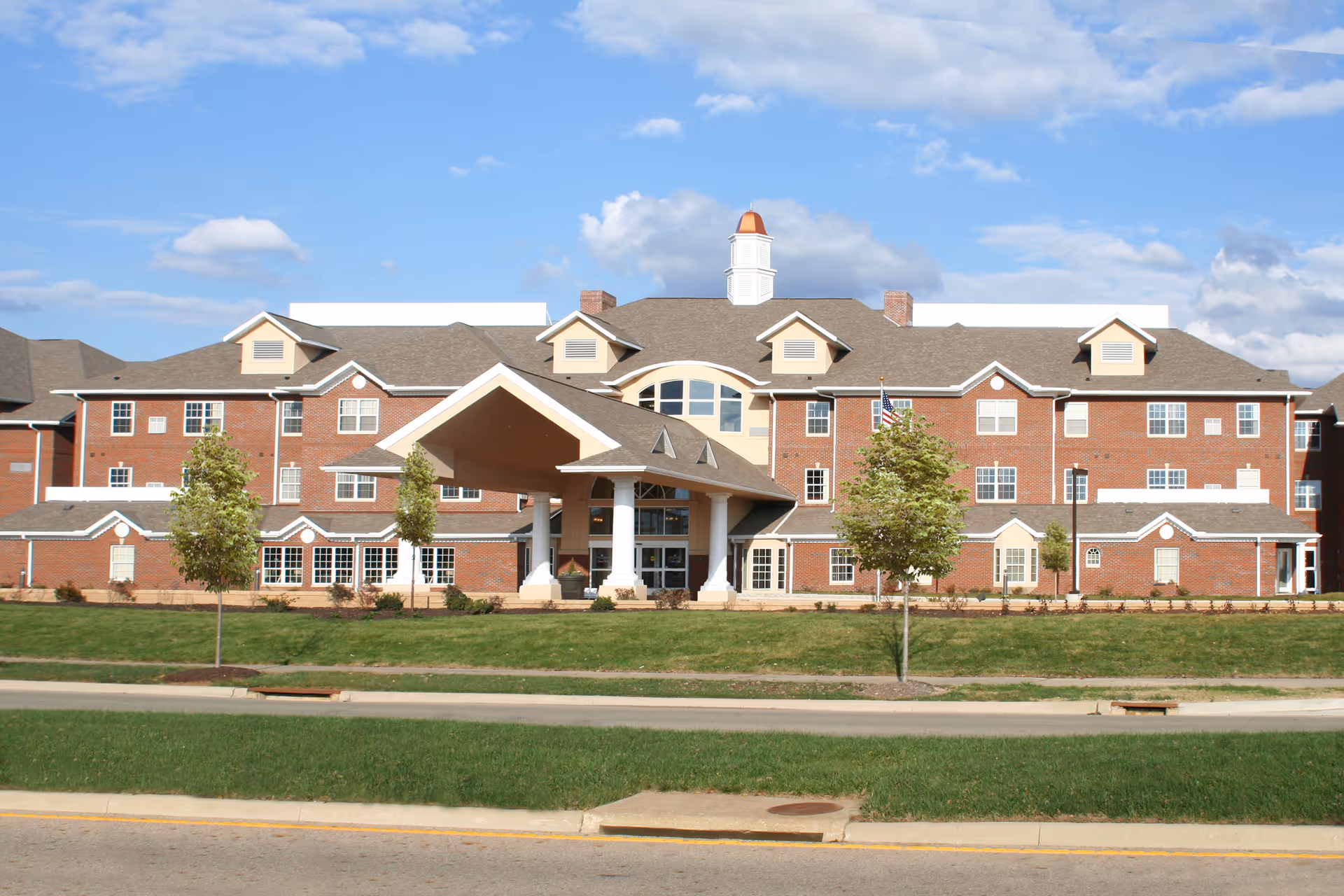 Front exterior view of a large brick building with multiple windows, a covered entrance supported by white columns, a cupola on the roof, and a well-maintained lawn with small trees in front under a partly cloudy blue sky.