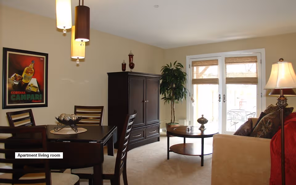A cozy apartment living room with a dining table and four chairs on the left, a dark wooden cabinet against the wall, a round coffee table in the center, a beige sofa with cushions on the right, a floor lamp beside the sofa, and large glass doors leading to an outdoor patio. A framed poster hangs on the wall above the dining table, and a tall potted plant is near the glass doors.