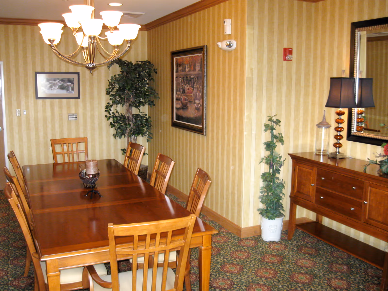 A dining room with a long wooden table surrounded by eight wooden chairs with cushioned seats. The walls have yellow striped wallpaper and are decorated with framed artwork. There is a chandelier with multiple lights hanging above the table. A wooden sideboard with drawers and a lamp with a dark shade is positioned against one wall, next to a tall potted plant. The floor is carpeted with a patterned design.