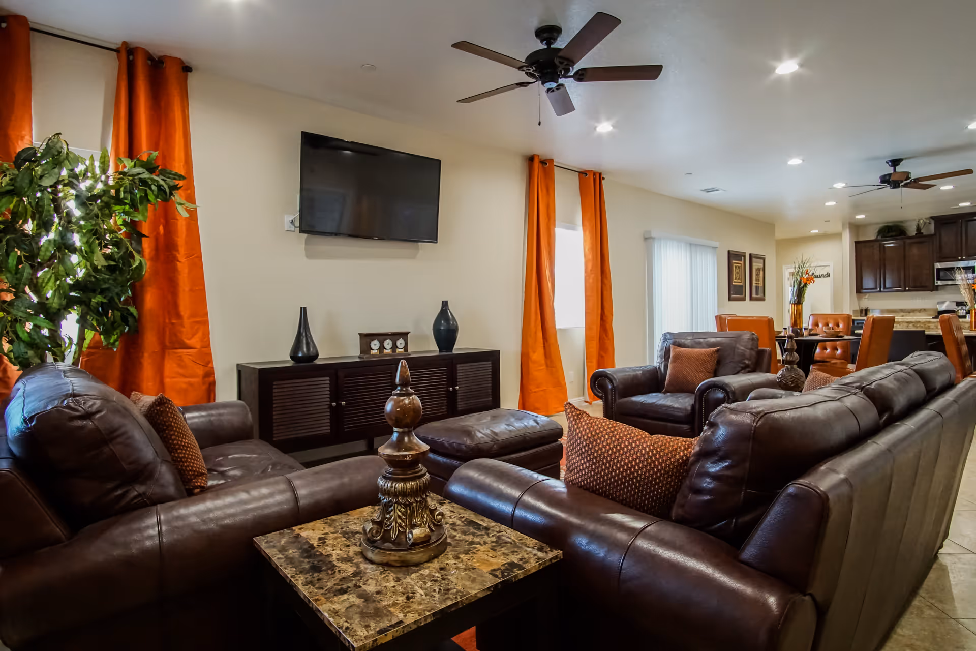 A cozy living room with dark brown leather sofas and armchairs arranged around a marble-top coffee table. The room features orange curtains on the windows, a flat-screen TV mounted on the wall, decorative vases on a wooden console, ceiling fans, and recessed lighting. In the background, a dining area with orange cushioned chairs and a kitchen with dark wood cabinets are visible.