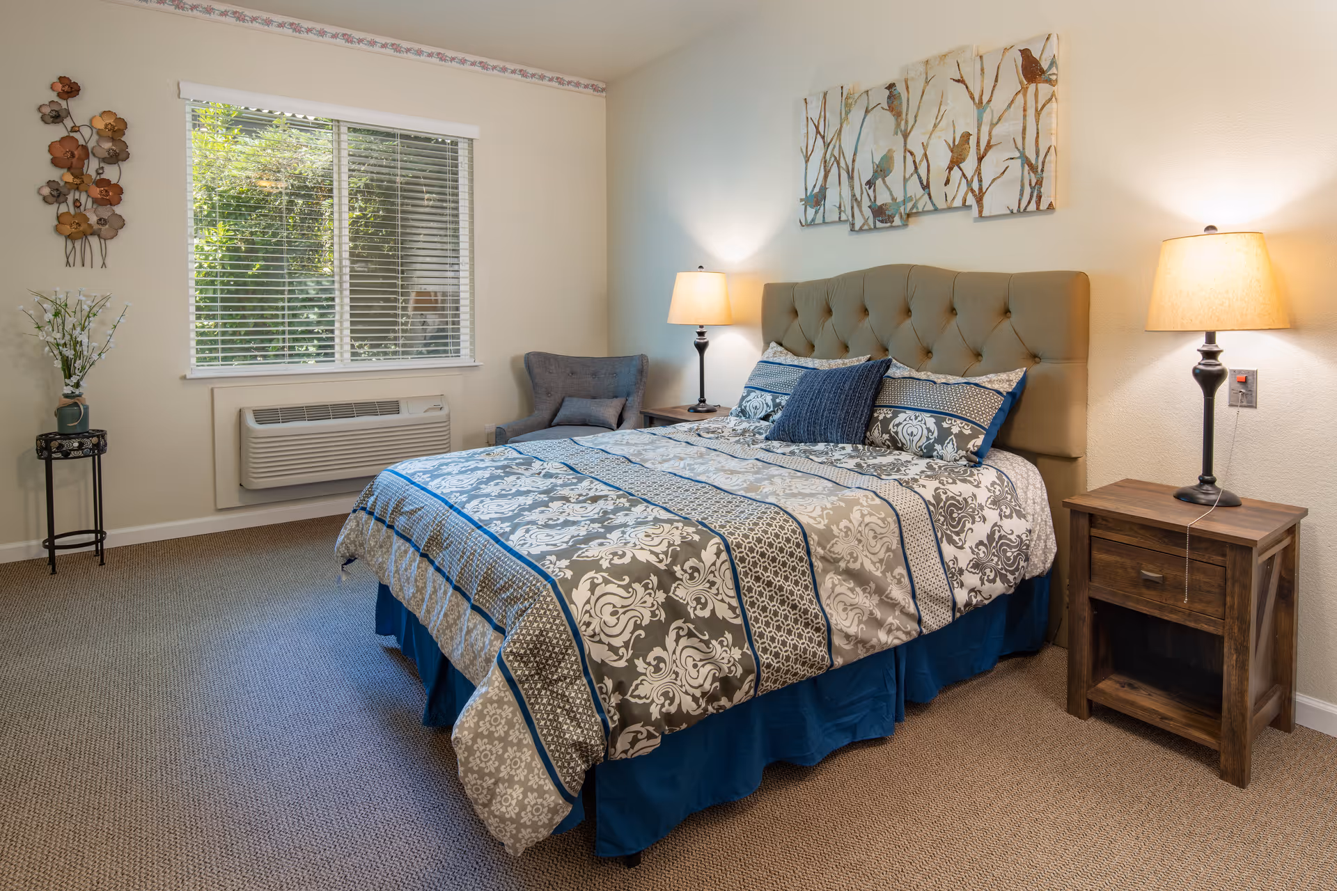 A neatly arranged bedroom with a bed topped by patterned bedding and a tufted headboard, flanked by nightstands with lamps, an armchair, and a window with blinds.