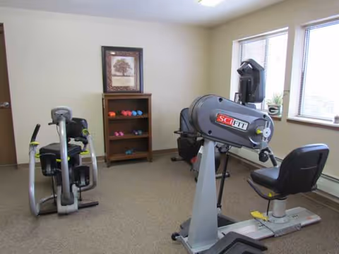 Sunlit interior exercise room with stationary bike, SciFit machine, and a weight shelf.
