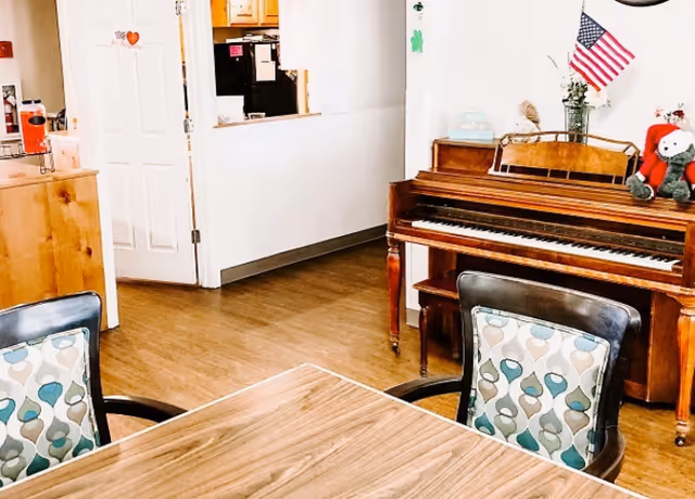 Communal room with a wooden piano against the wall, patterned chairs around a wood table, and an open doorway to a kitchen area.