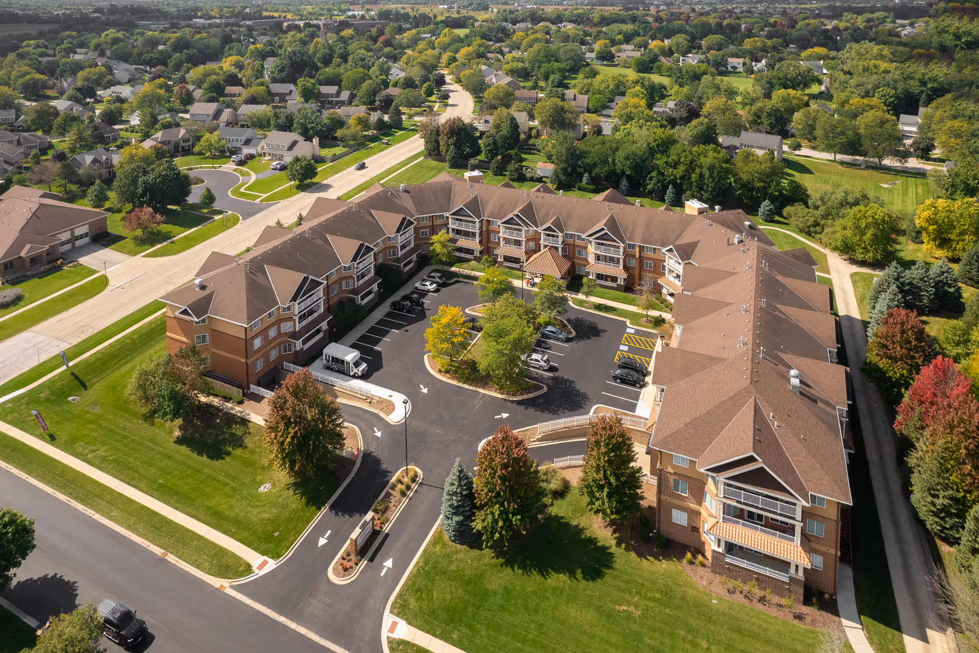 Aerial view of a multi-story senior living complex arranged around a central parking courtyard amid lawns and surrounding suburban houses.
