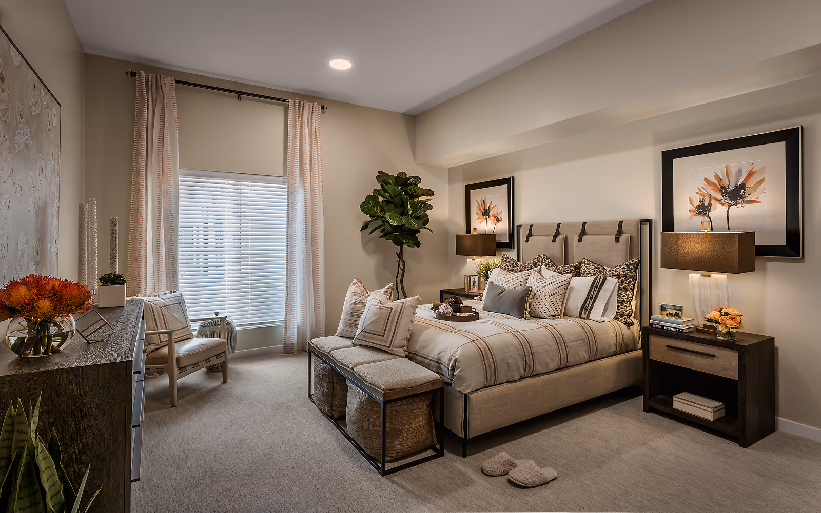 A cozy, well-decorated bedroom with a large bed featuring multiple pillows and a beige plaid comforter. There is a cushioned bench at the foot of the bed with storage baskets underneath. The room has beige walls, a large window with blinds and light pink curtains, a wooden dresser with decorative items, a comfortable chair, two framed floral artworks above the bed, two bedside tables with lamps, and a tall green plant in the corner. A pair of slippers is placed on the carpeted floor near the bed.