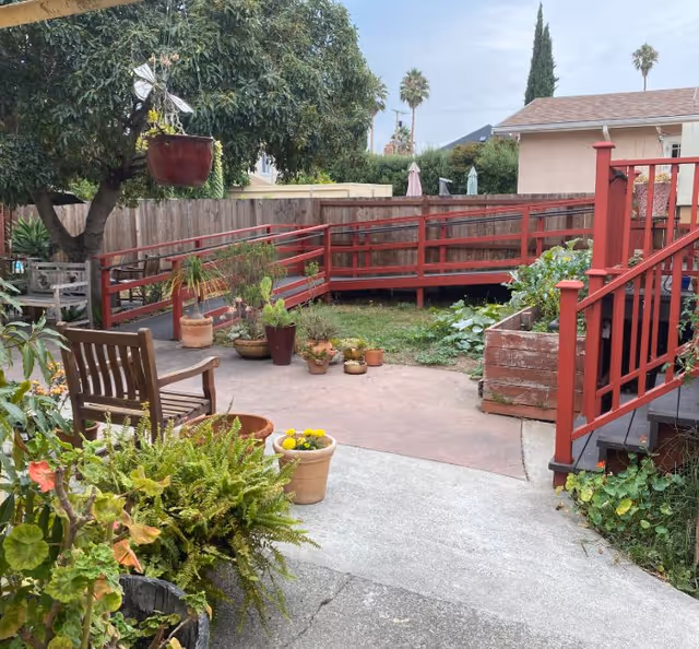 Outdoor garden area with various potted plants, a wooden bench, a red wooden ramp and railing, and a tree with a hanging plant pot. There is a wooden fence in the background and a house with a tiled roof visible beyond the fence.