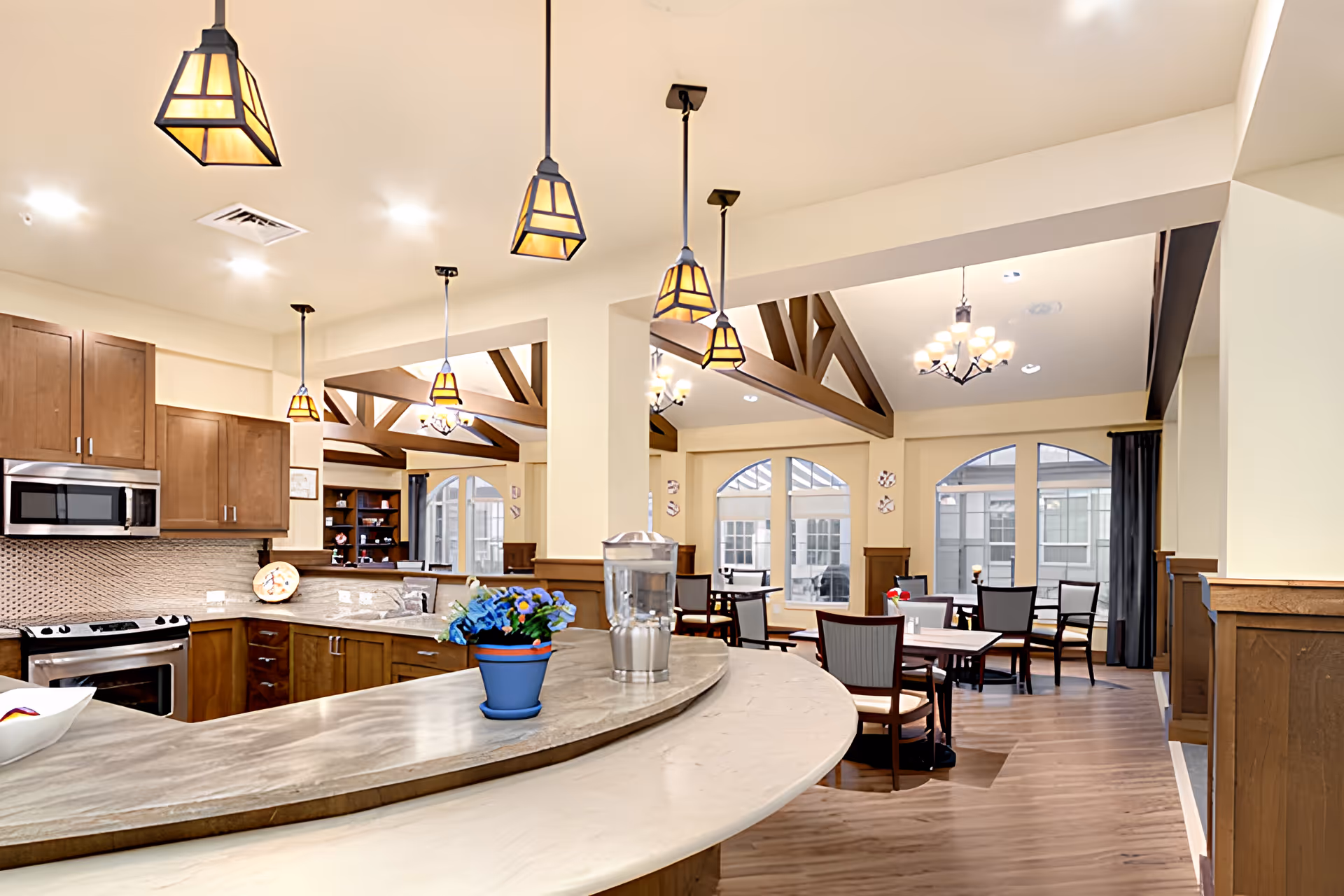Interior view of a bright and spacious dining area and kitchen in a senior living facility. The kitchen features wooden cabinets, a stainless steel oven and microwave, and a curved countertop with a blue potted plant and a water pitcher. The dining area has multiple tables and chairs, large windows with arched tops, wooden beams on the ceiling, and pendant lights hanging above the kitchen counter.
