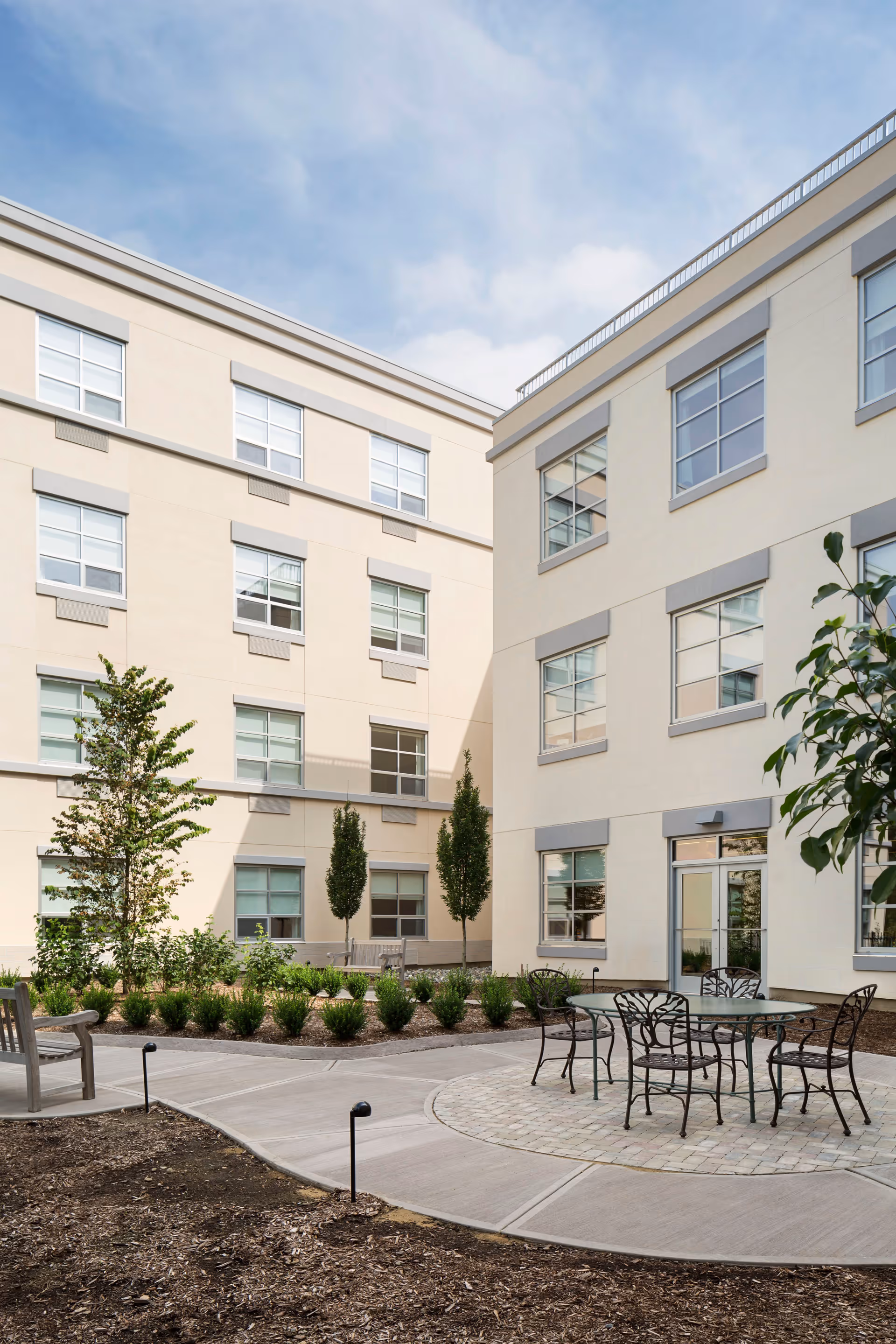 Outdoor courtyard area of a senior living facility with beige multi-story buildings surrounding a paved circular patio. The patio has a glass-top table with four metal chairs. There are small trees, shrubs, and benches along the walkway, under a partly cloudy sky.
