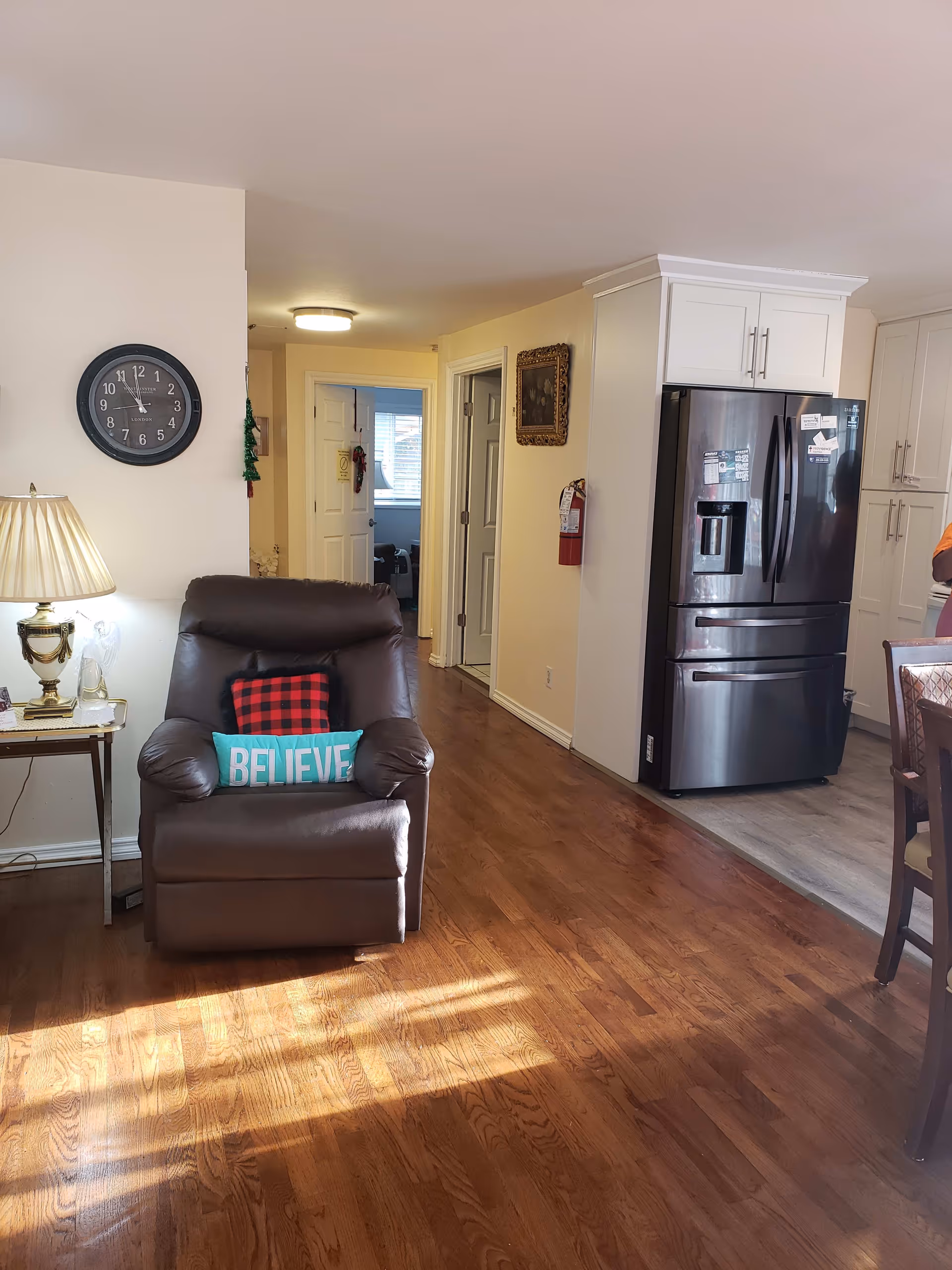 Interior view of a living space showing a brown leather recliner chair with two decorative pillows, one red and black checkered and one turquoise with the word 'BELIEVE'. To the left of the chair is a side table with a lamp and a glass angel figurine. A wall clock is mounted above the chair. In the background, there is a hallway with open doors and a kitchen area with a stainless steel refrigerator and white cabinets.