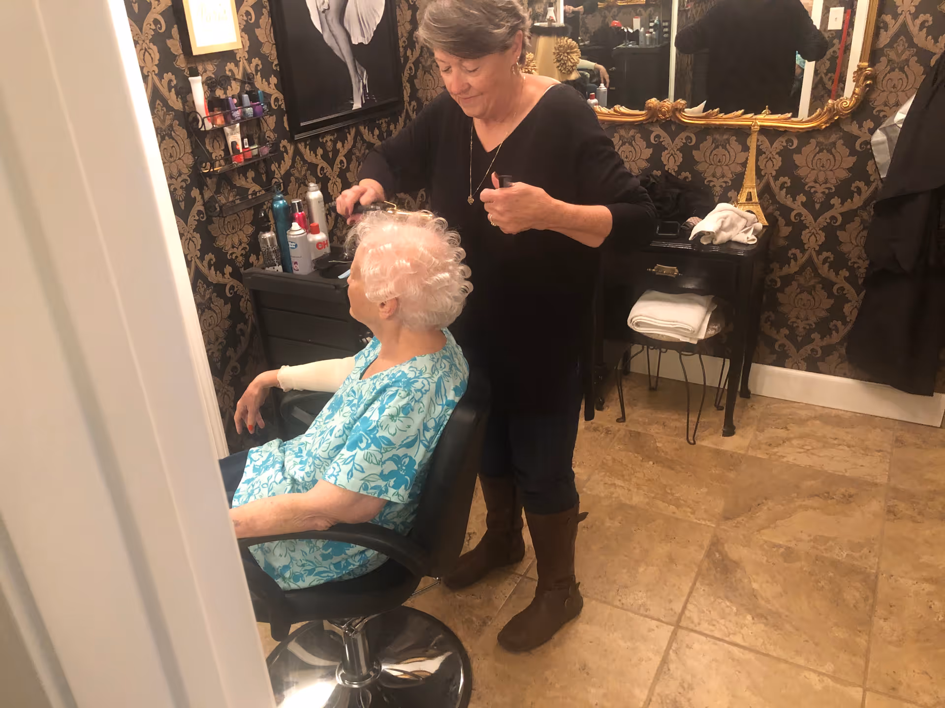 An elderly woman with white curly hair sits in a salon chair while another woman styles her hair in a room with patterned wallpaper, a large ornate mirror, and various hair care products on a shelf.