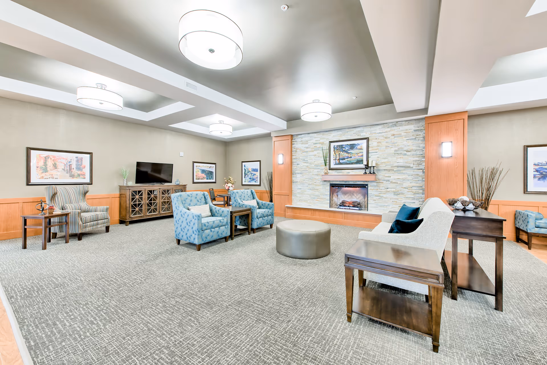 A spacious and well-lit living room area with modern ceiling lights, a stone fireplace with a painting above it, multiple comfortable chairs and sofas arranged around a central ottoman, side tables, and a TV on a wooden cabinet. The room has a neutral color palette with light walls and carpeted flooring.