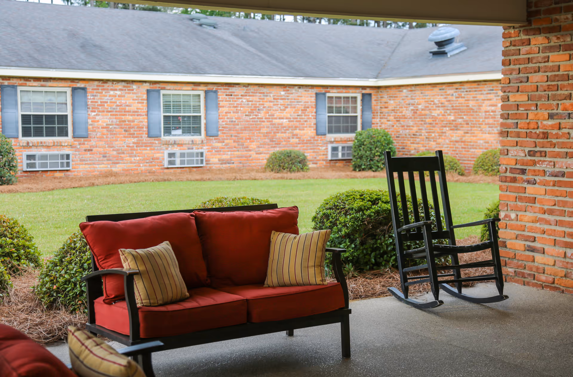 Covered patio with a red-cushioned loveseat and a black rocking chair overlooking a grassy courtyard and a brick building.