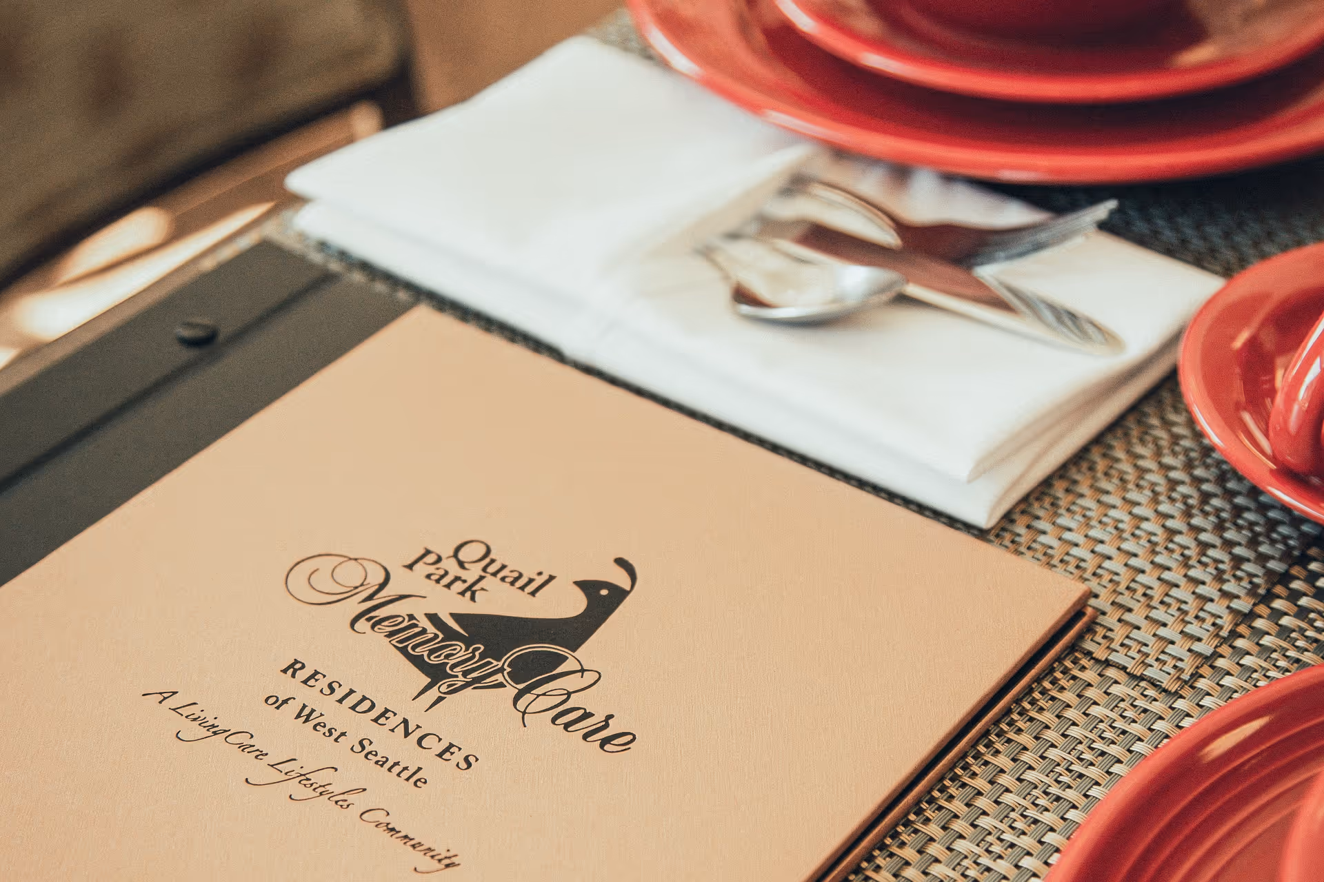 A Quail Park Memory Care brochure on a table next to a folded napkin, silverware, and red dishes.