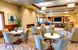 Bright communal lounge with round tables and upholstered chairs, a fireplace, TV, and decorative artwork.