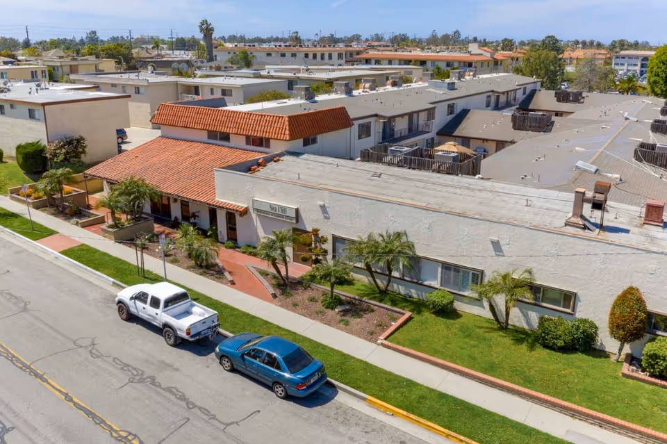 Aerial view of the Sea Cliff Assisted Living building front with a tiled roof, landscaped lawn, walkways, and two cars parked on the street.