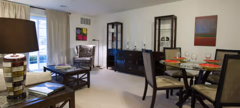 A well-lit living and dining area featuring a glass-top dining table set with four chairs and red placemats. The living area includes a coffee table, an armchair with a patterned fabric, a side table with a lamp, and a large window with beige curtains. Against the wall are two dark wood display cabinets and a sideboard with decorative items and artwork hanging above.
