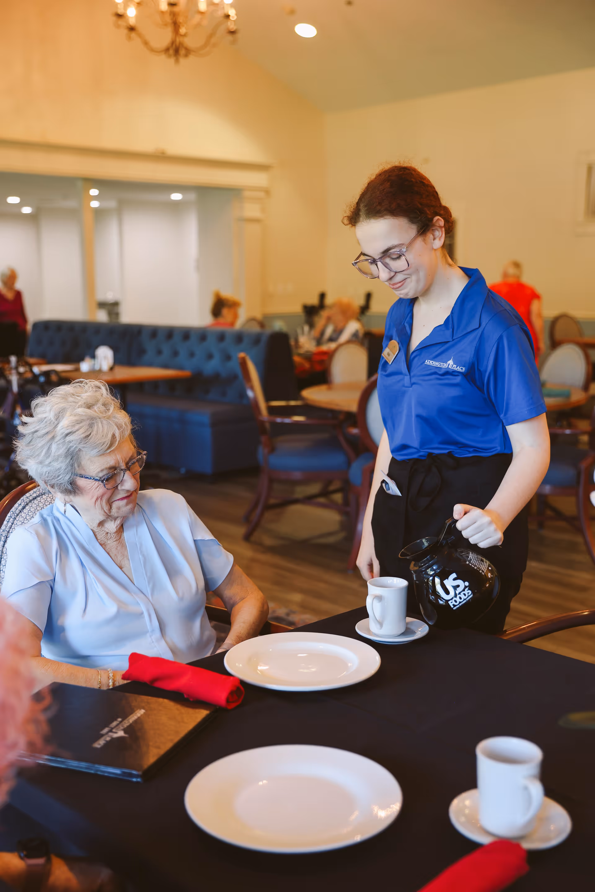A young female staff member in a blue Addington Place polo shirt is pouring coffee into a cup for an elderly woman seated at a dining table in a senior living facility dining room. The elderly woman is wearing glasses and a light blue blouse. The table is set with white plates, red napkins, and coffee cups. Other residents and tables are visible in the background.
