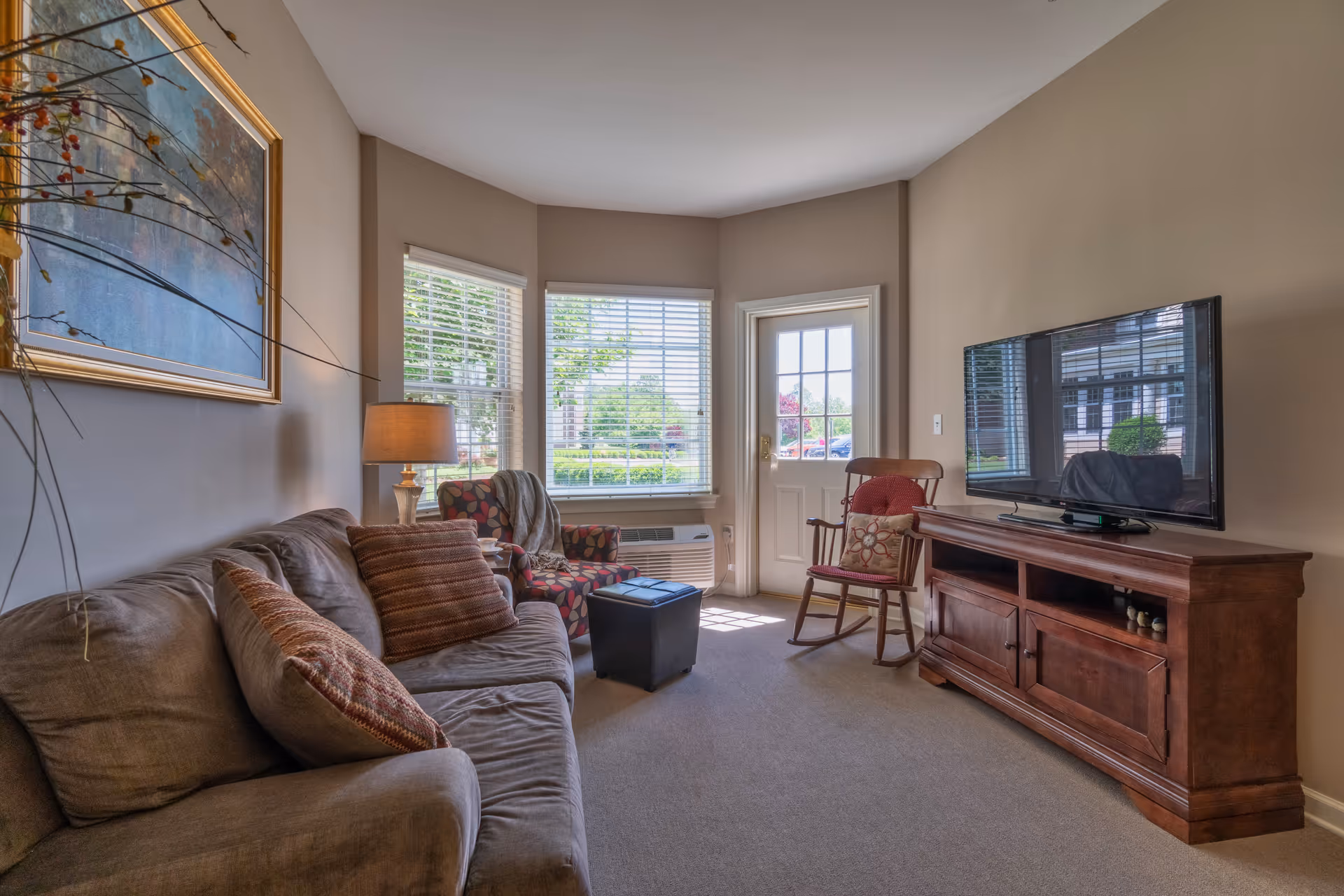 A cozy living room with a brown sofa adorned with patterned pillows, a colorful armchair with a throw blanket, a wooden rocking chair with a cushion, a wooden TV stand with a flat-screen television, and a lamp on a side table. The room has beige walls, carpeted floor, and windows with white blinds letting in natural light. A door with glass panels leads outside.