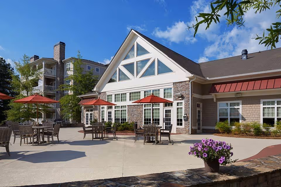 Outdoor patio area of The Lodge at Bridgemill featuring multiple wooden tables and chairs with red umbrellas, a stone and siding building with large windows and a triangular glass section at the roof peak, and a potted plant with purple flowers in the foreground under a blue sky with some clouds.