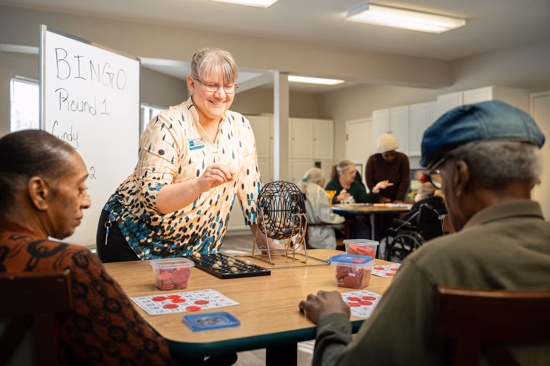 A group of elderly people playing bingo in a well-lit room at an assisted living facility. A smiling woman is operating the bingo cage while two seniors sit at the table with bingo cards and markers. Other residents are visible in the background, engaged in activities around another table. A whiteboard behind the woman reads 'BINGO Round 1 Cindy'.