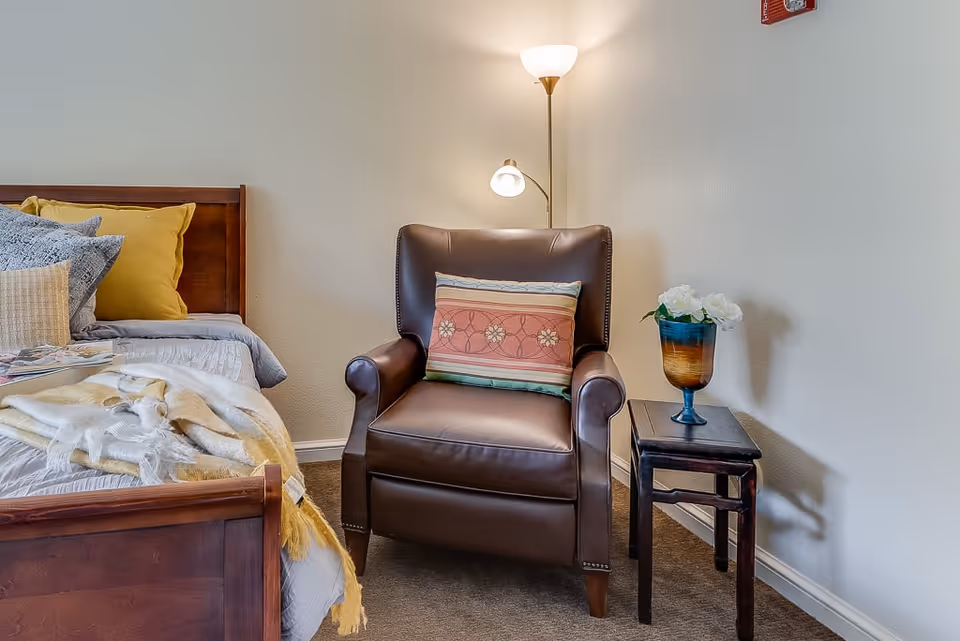 A cozy corner of a bedroom featuring a wooden bed with multiple pillows and a blanket, a brown leather armchair with a decorative pillow, a small wooden side table with a vase of white flowers, and a floor lamp providing warm light against a plain wall.