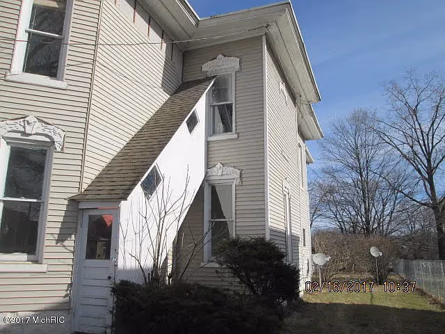 Exterior front corner of a beige two-story house with a small angled entryway, decorative window trim, shrubs, and bare trees.