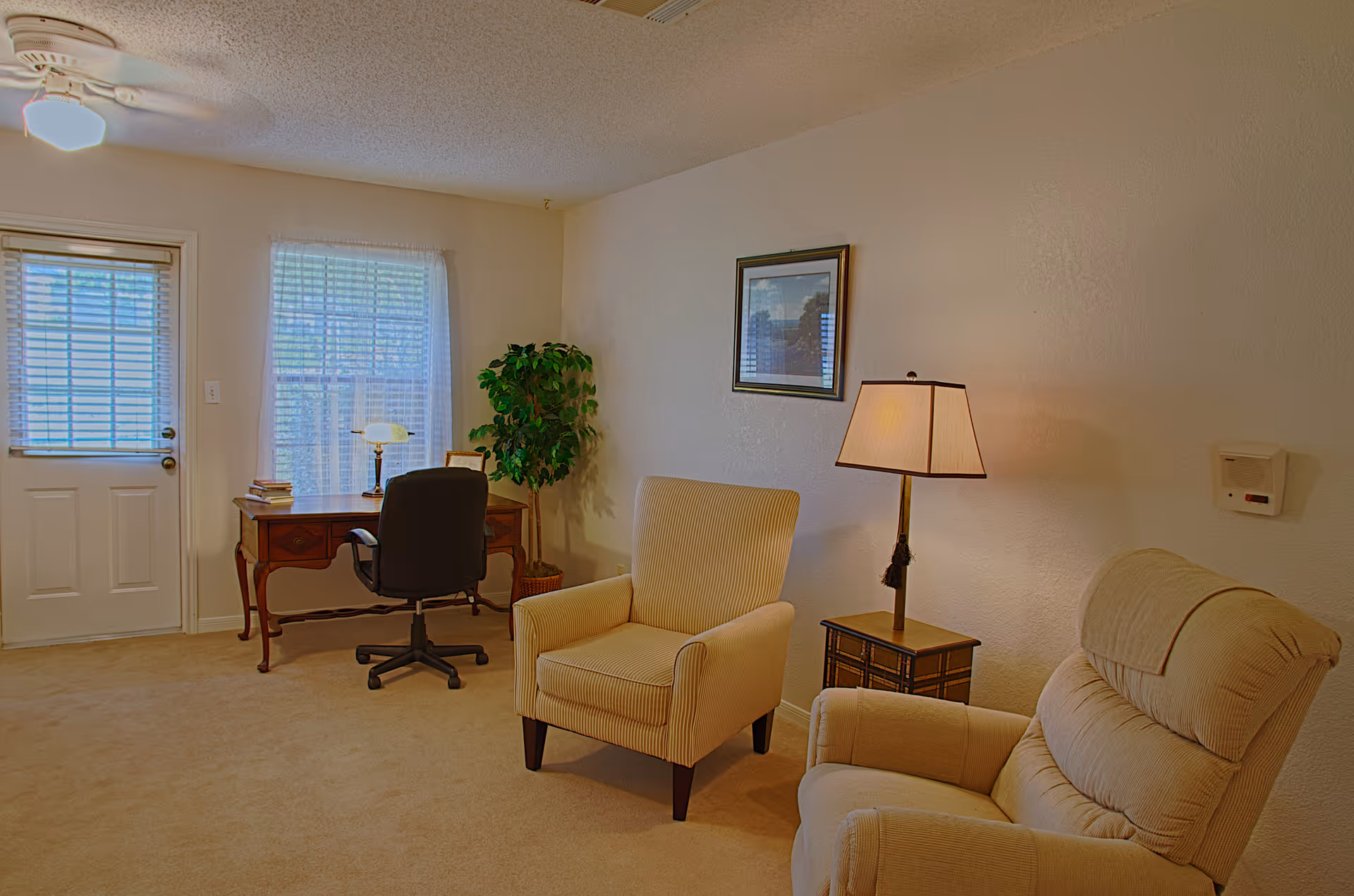 A cozy interior room with beige carpet and walls, featuring a wooden desk with a black office chair, a table lamp, and a stack of books near a window with blinds and sheer curtains. There are two upholstered armchairs, one beige and one light tan, positioned near a small side table with a lamp. A framed picture hangs on the wall above the beige armchair, and a potted plant is placed in the corner near the desk.