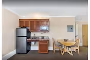 Small kitchenette area with a refrigerator, microwave, sink, and wooden cabinets on the left side. To the right, there is a round wooden dining table with four chairs. The walls are beige, and the floor is carpeted. A door and an electrical panel are visible in the background.