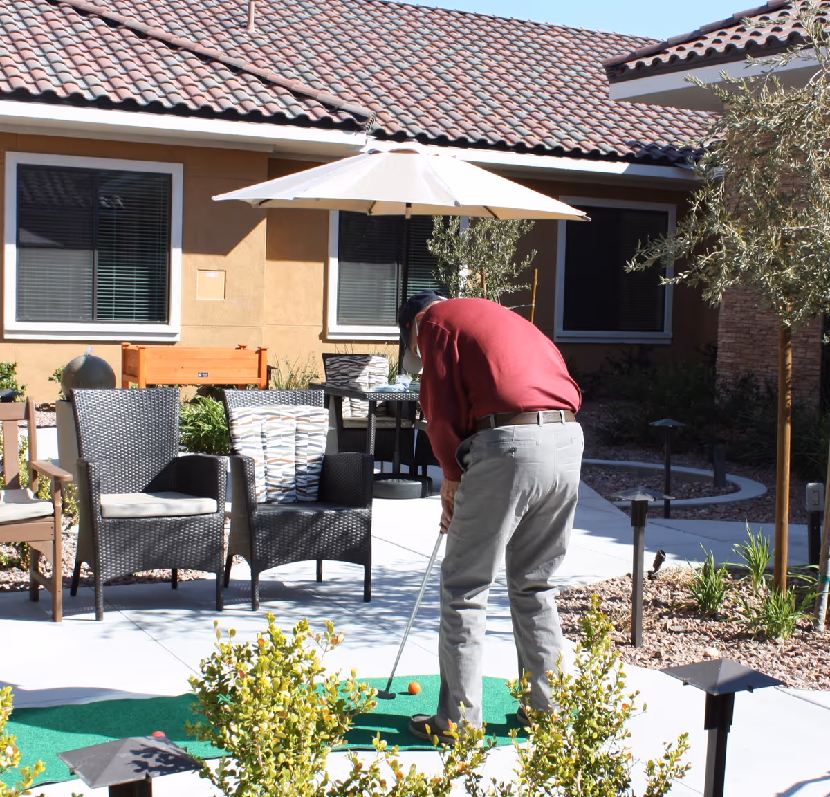 A person practices putting on a small outdoor putting green in a courtyard with patio furniture and an umbrella.