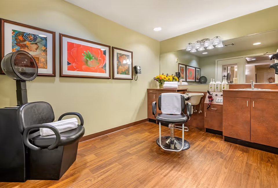 Interior view of a hair salon area with a black salon chair and hair dryer on the left, a styling chair with a white towel draped over it in the center, wooden cabinets with a sink and hair care products on the right, three framed pictures on the wall, and a large mirror reflecting the room.