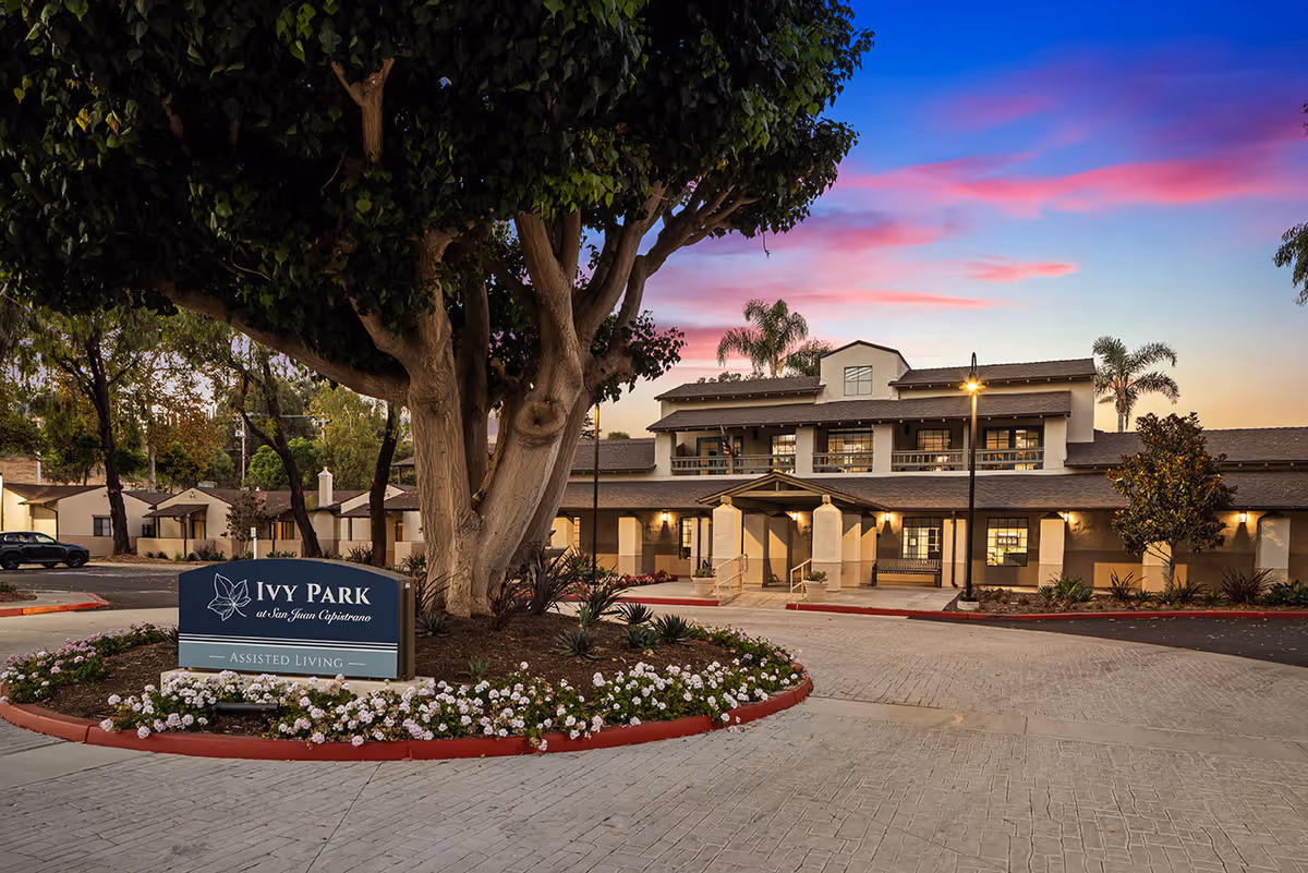 Exterior view of Ivy Park at San Juan Capistrano assisted living facility during sunset, showing a large building with multiple windows, a covered entrance, palm trees, and a landscaped roundabout with flowers and a sign displaying the facility's name.