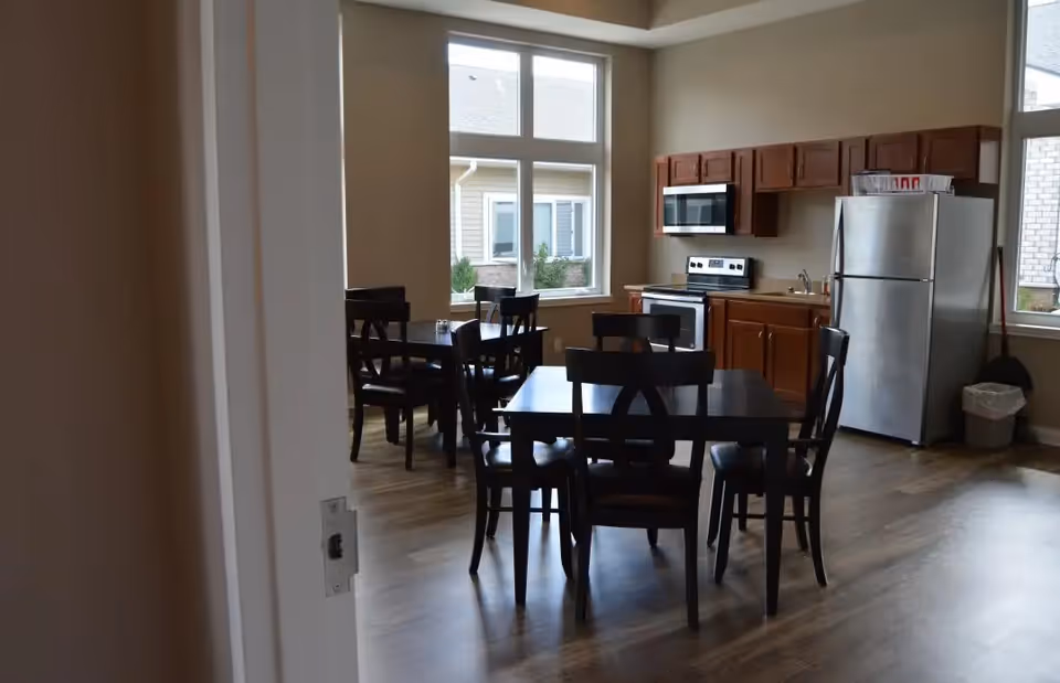 Interior view of a dining area with dark wooden tables and chairs, a kitchen area with wooden cabinets, a stainless steel refrigerator, stove, and microwave. Large windows allow natural light into the room.
