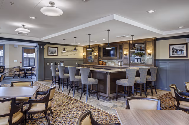 A spacious and well-lit dining area with a large central bar surrounded by high chairs. The bar has wooden cabinetry, a mounted TV, and pendant lighting. Several tables and chairs are arranged around the room, with patterned carpet and tiled flooring near the bar. The walls are decorated with framed pictures and a wall quote.