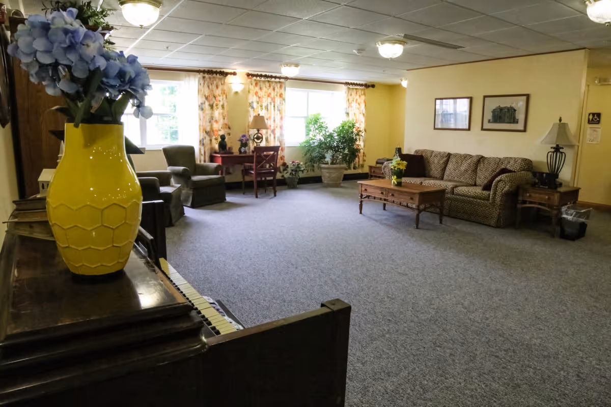 A spacious living room area in a senior living facility with a patterned sofa, wooden coffee table with a small flower arrangement, two armchairs, a wooden desk with a lamp, and large windows with floral curtains. A yellow vase with blue flowers is placed on a piano in the foreground. The room has carpeted flooring and soft ceiling lights.