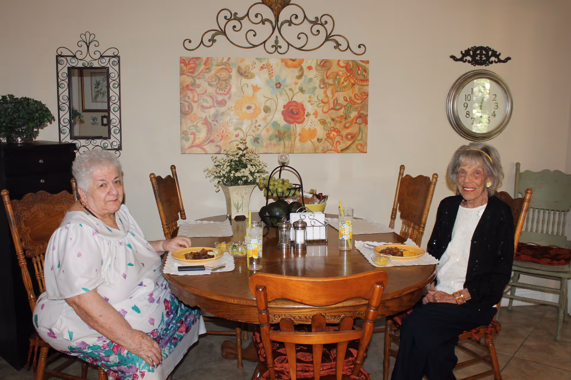Two elderly women sitting at a round wooden dining table with plates of food and glasses of water with straws. The room has a floral painting on the wall, a decorative clock, a mirror, and a vase with flowers on the table.
