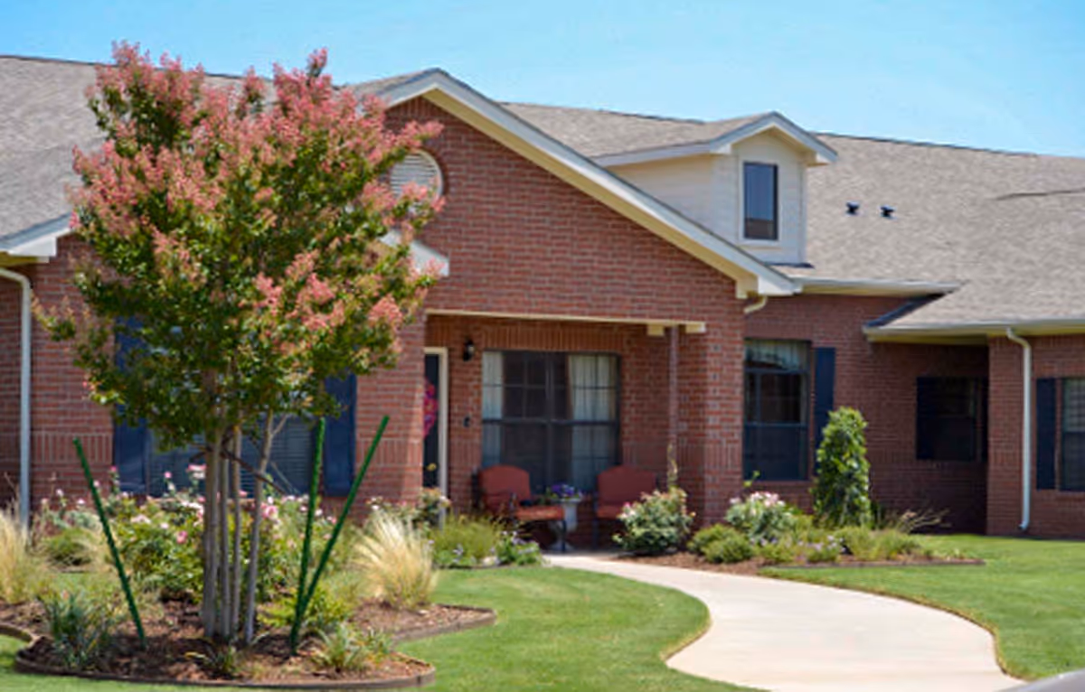Exterior view of a single-story brick building with a gabled roof, featuring a small porch with two chairs and a table. The building is surrounded by a well-maintained lawn, landscaped flower beds, and a tree with pink blossoms. A curved concrete walkway leads to the entrance.