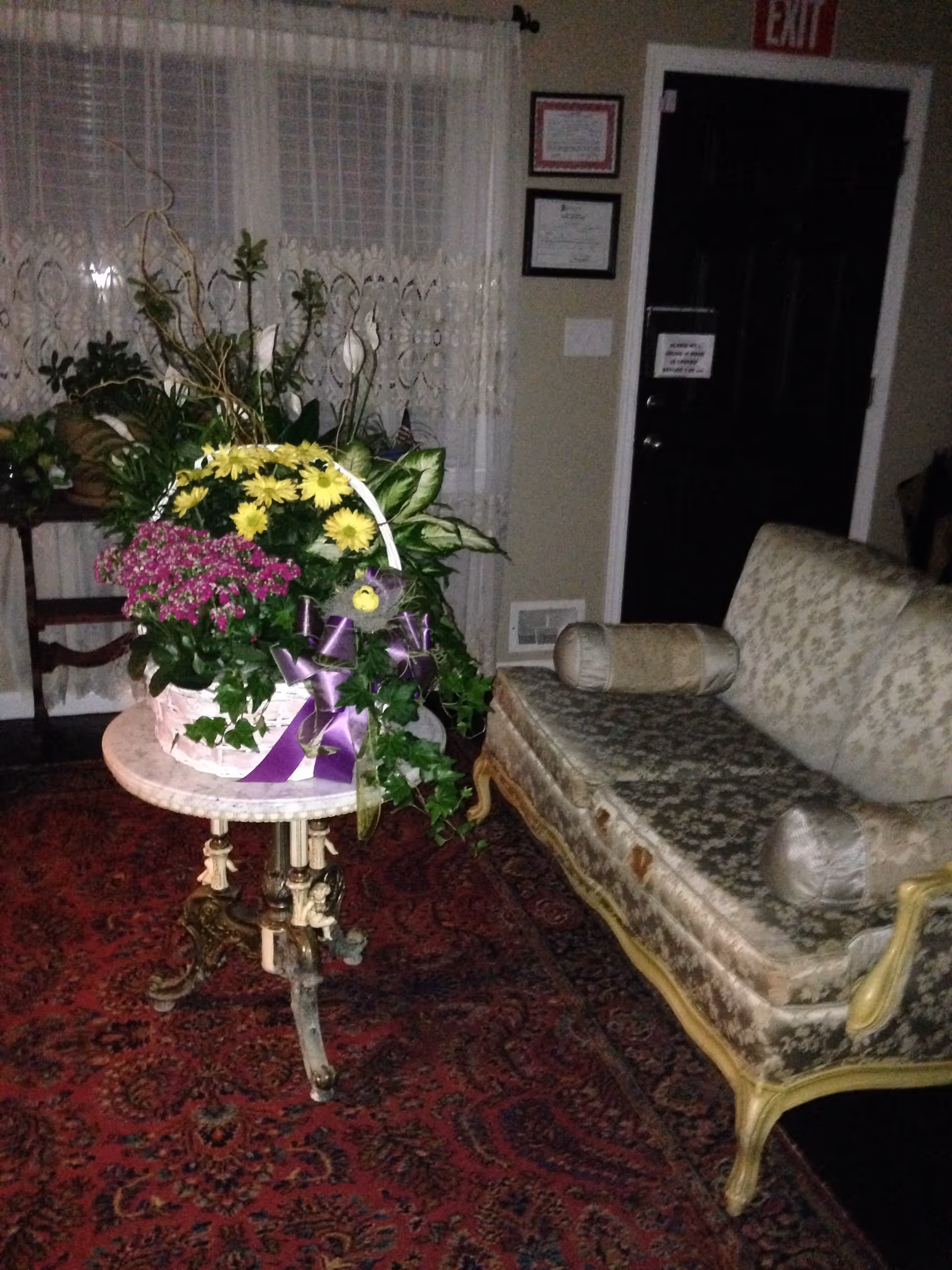 A cozy interior room with a vintage floral-patterned sofa with two cylindrical pillows, a small ornate round table with a large flower arrangement featuring yellow and purple flowers, and a red patterned carpet. The background shows a window with lace curtains, framed certificates on the wall, and a black door with an exit sign above it.