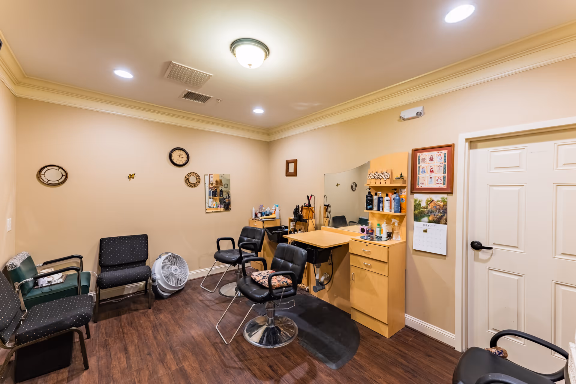 Interior of a senior living facility's hair salon room with beige walls, wood flooring, a styling station with mirror and hair care products, several chairs including salon chairs and waiting chairs, a wall clock, and a calendar on the wall.