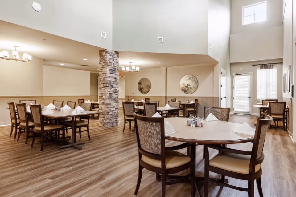 A spacious dining room with multiple wooden tables and chairs arranged neatly. Each table is set with white folded napkins and condiment holders. The room features a stone pillar, light wood flooring, soft beige walls, and two round decorative wall hangings. Large windows and a glass door allow natural light to brighten the space.