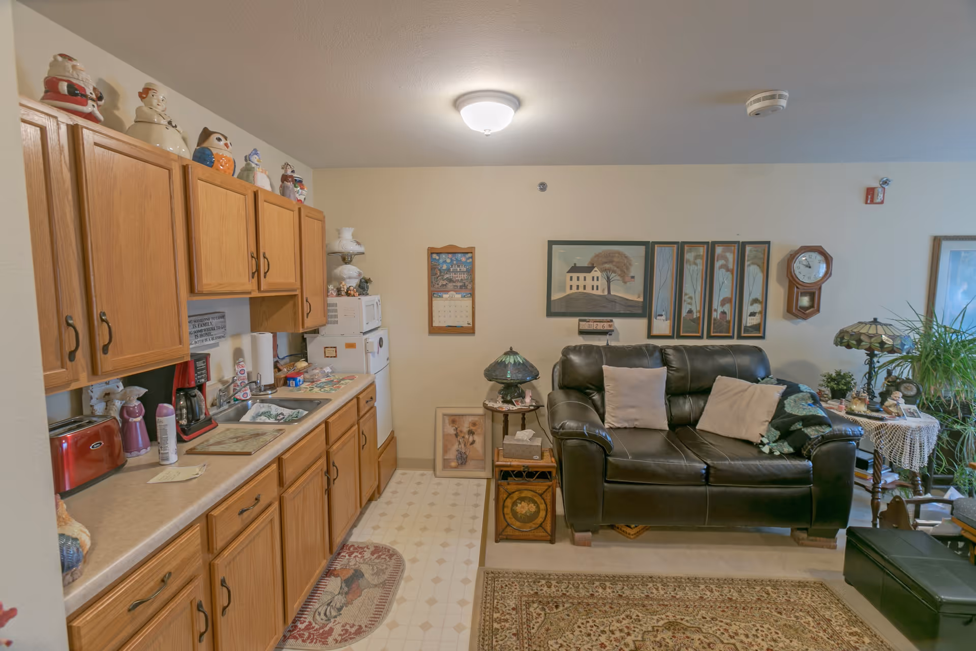 Interior view of a senior living facility room featuring a kitchenette with wooden cabinets, a countertop with a sink, coffee maker, toaster, and various decorative items. Adjacent to the kitchenette is a living area with a black leather loveseat, two beige pillows, side tables with lamps, wall art, a clock, and plants. The floor has a patterned rug and linoleum tiles.