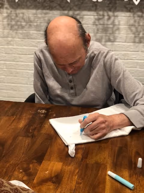 An elderly man with a bald head and gray long-sleeve shirt is sitting at a wooden table, focused on drawing or coloring on a white cloth with a blue marker. There are additional markers on the table and a white brick wall in the background.